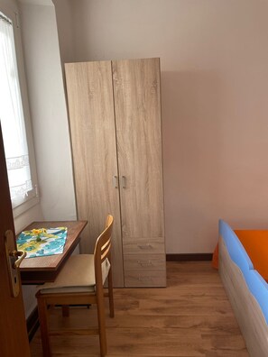 Una habitación con un armario de madera, una mesita con mantel floral, una silla de madera y una cama con cabecera naranja y azul.