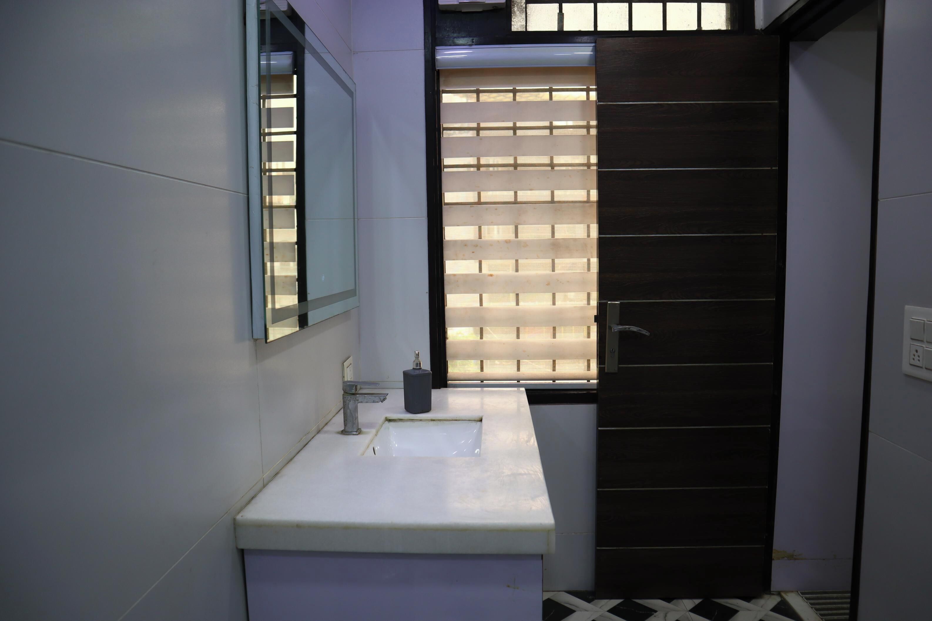 Un baño con un lavamanos blanco, un espejo, una ventana con persianas y una puerta oscura.