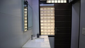 Un baño con un lavamanos blanco, un espejo, una ventana con persianas y una puerta oscura.