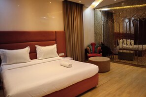 A hotel room with a large bed, a small round ottoman, a seating area with chairs, and a view through a glass partition. - The Royal Suites (New Delhi)