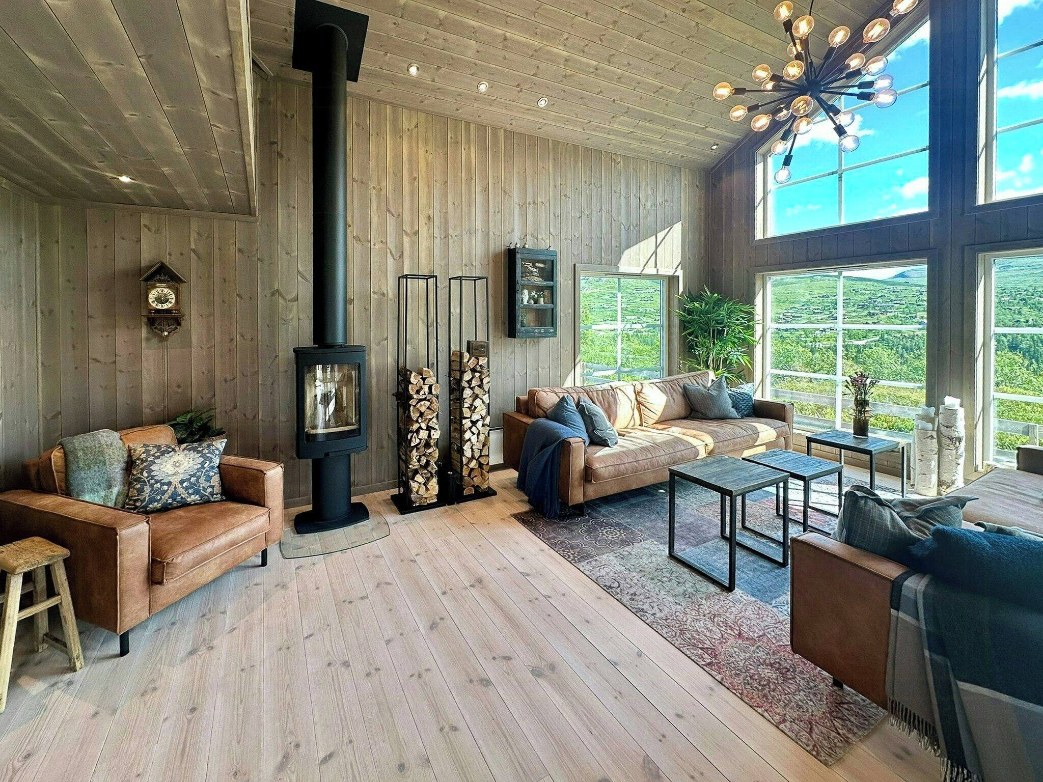 A cozy living room with a fireplace, wooden flooring, and large windows offering a view of the outdoors.