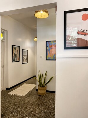 A hallway with a patterned carpet, a potted plant, framed artwork on the walls, and a door on the left.