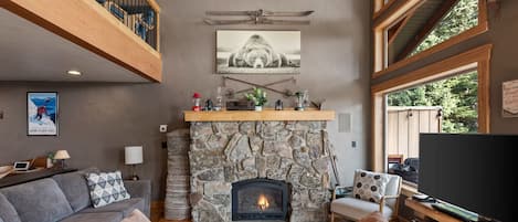 A cozy living room with a stone fireplace, a comfortable sofa, a television, and a large window offering a view of the outdoors.