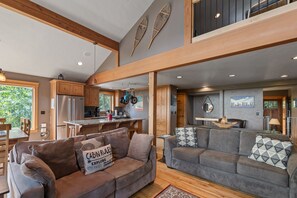 A spacious living area with a high ceiling, wooden beams, and a kitchen in the background. - Snowplow Home | Hot Tub + Dog Friendly (Sandpoint)