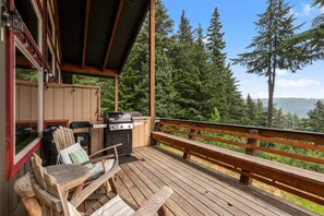 A wooden deck with a grill, chairs, and a view of a forest. - Snowplow Home | Hot Tub + Dog Friendly (Sandpoint)
