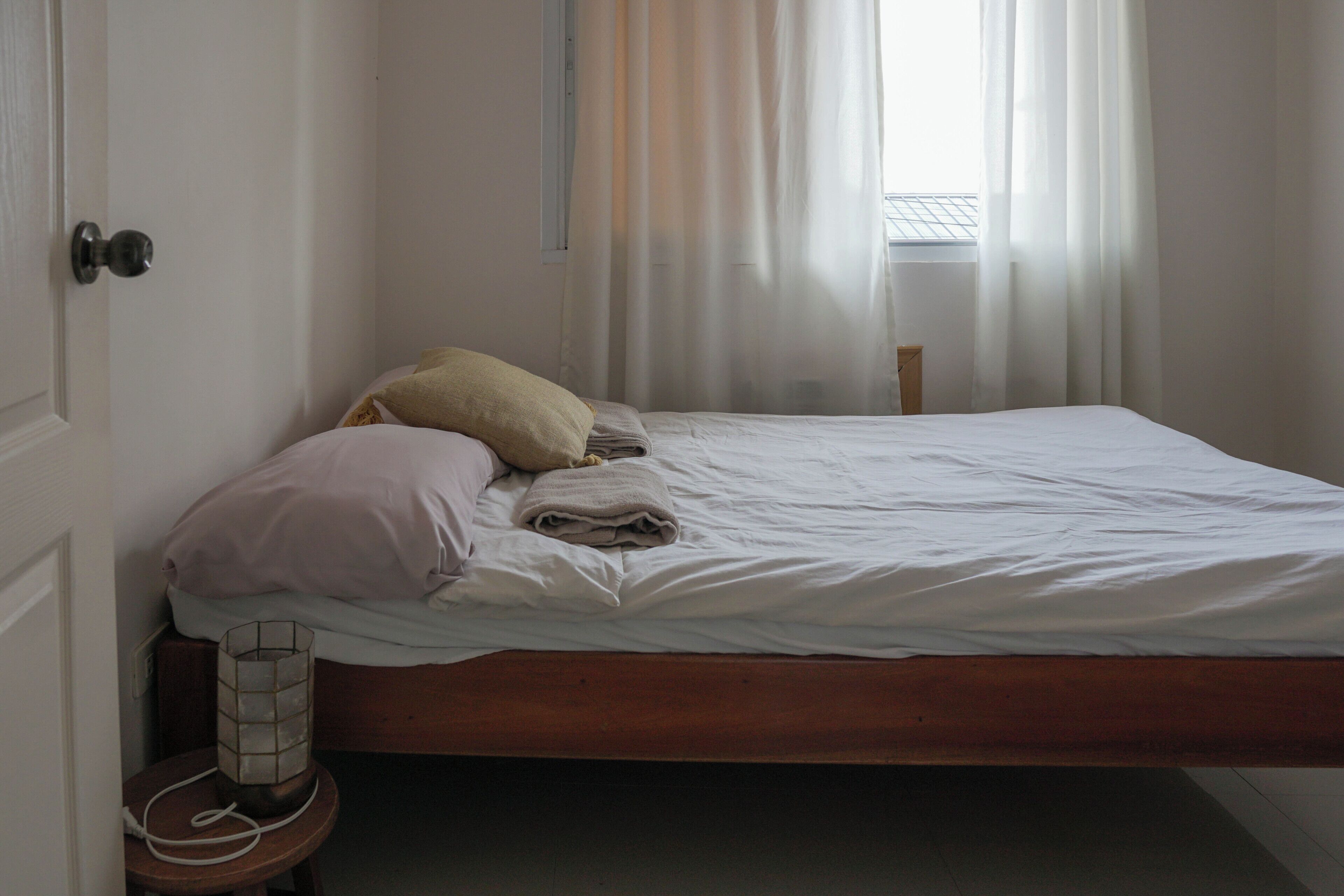 A neatly made bed with pillows and a blanket, a bedside table with a lamp, and a window with sheer curtains.