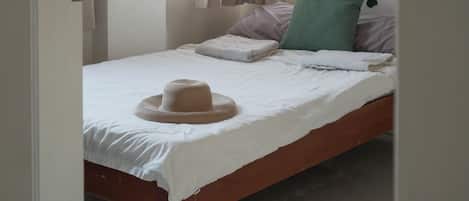 Una cama individual con sábana blanca, un sombrero tejido y una almohada verde.