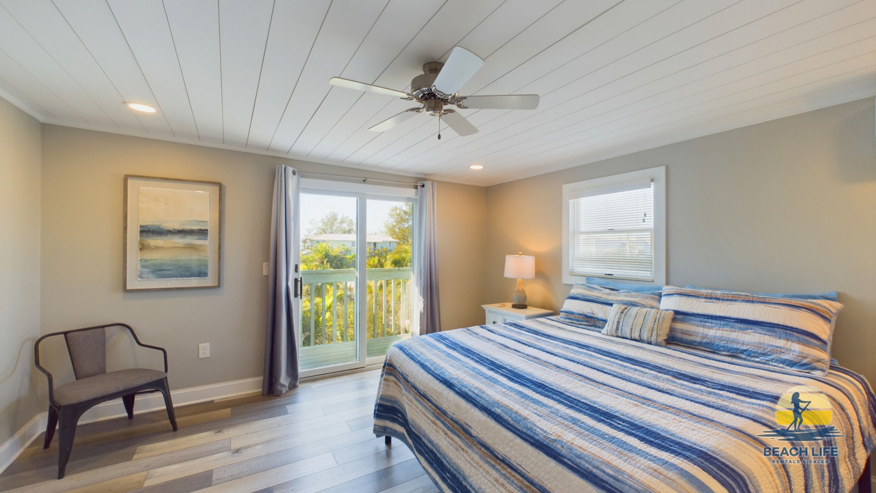 A bedroom with a bed, a chair, a ceiling fan, a window, and a door leading to a balcony.