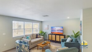 A living room with a sofa, armchairs, a coffee table, a TV, and a window with blinds. - Castaway Cottage Beautiful Pool Spa Steps to Sand Restaurants Golf Cart Included (Holmes Beach)
