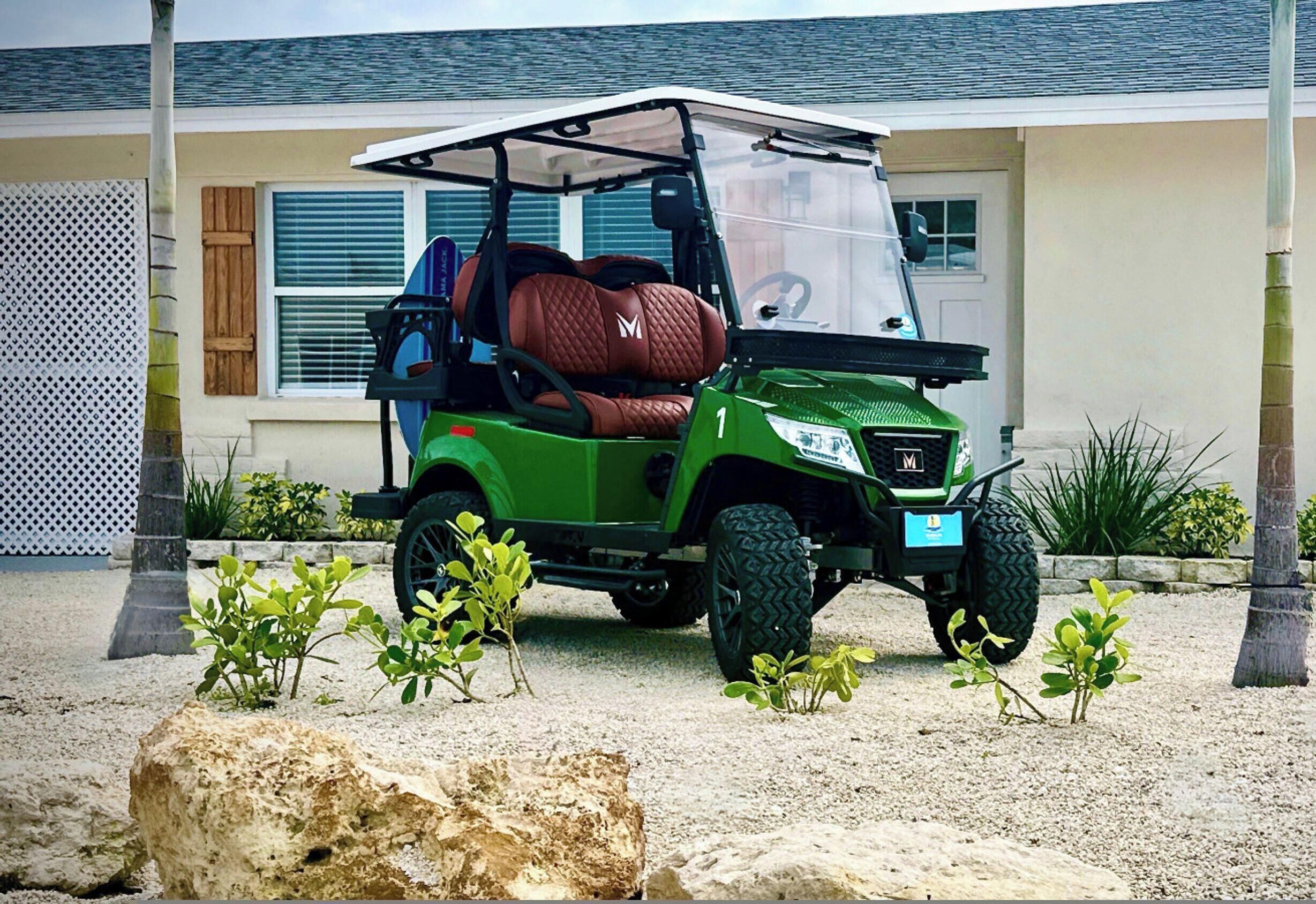 A green golf cart parked in front of a house with a white fence, a window with blinds, and a small garden with plants.
