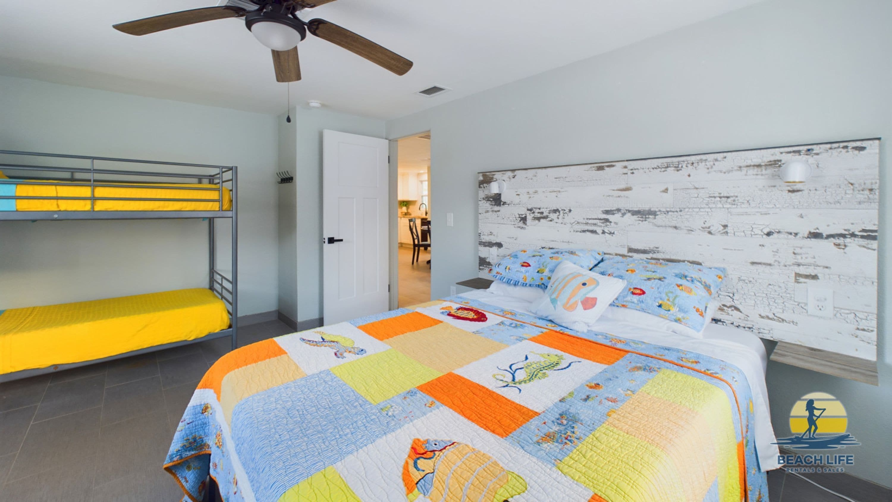 Une chambre avec des lits superposés, un lit recouvert d’une couette aux couleurs vives, un ventilateur de plafond et une porte blanche donnant sur une salle de bain.
