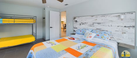 Un dormitorio con literas, una cama con colcha de colores, un ventilador de techo y una puerta blanca que da al baño.