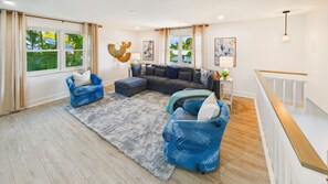 Ein geräumiges Wohnzimmer mit einem großen Ecksofa, zwei blauen Sesseln, einem grauen Shaggy-Teppich und Blick ins Grüne.