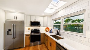 A modern kitchen with stainless steel appliances, wooden cabinets, and a large window offering a view of greenery. - Twin Tides Large Home Heated Pool Spa Putting Green Golf Cart Tiki Bar Ping Pong Sleeps 14 (Holmes Beach)