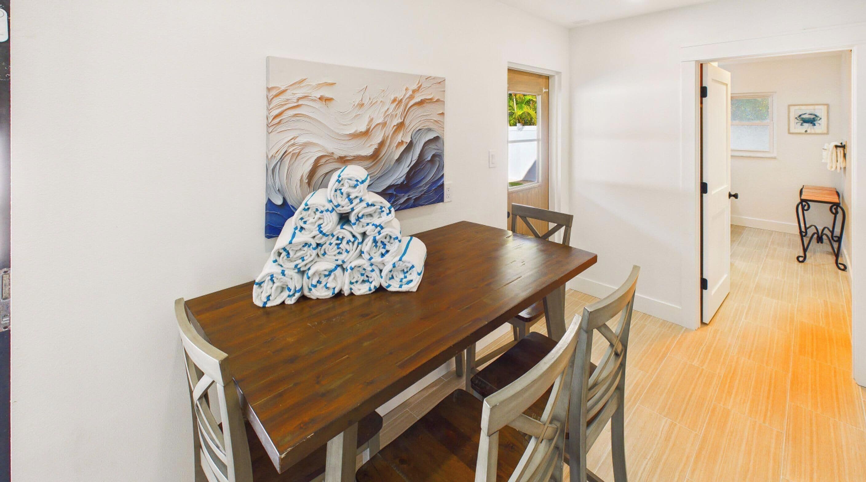 A dining area with a wooden table and chairs, a painting on the wall, and a view of the outdoors through an open door.