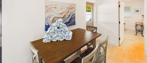 A dining area with a wooden table and chairs, a painting on the wall, and a view of the outdoors through an open door.