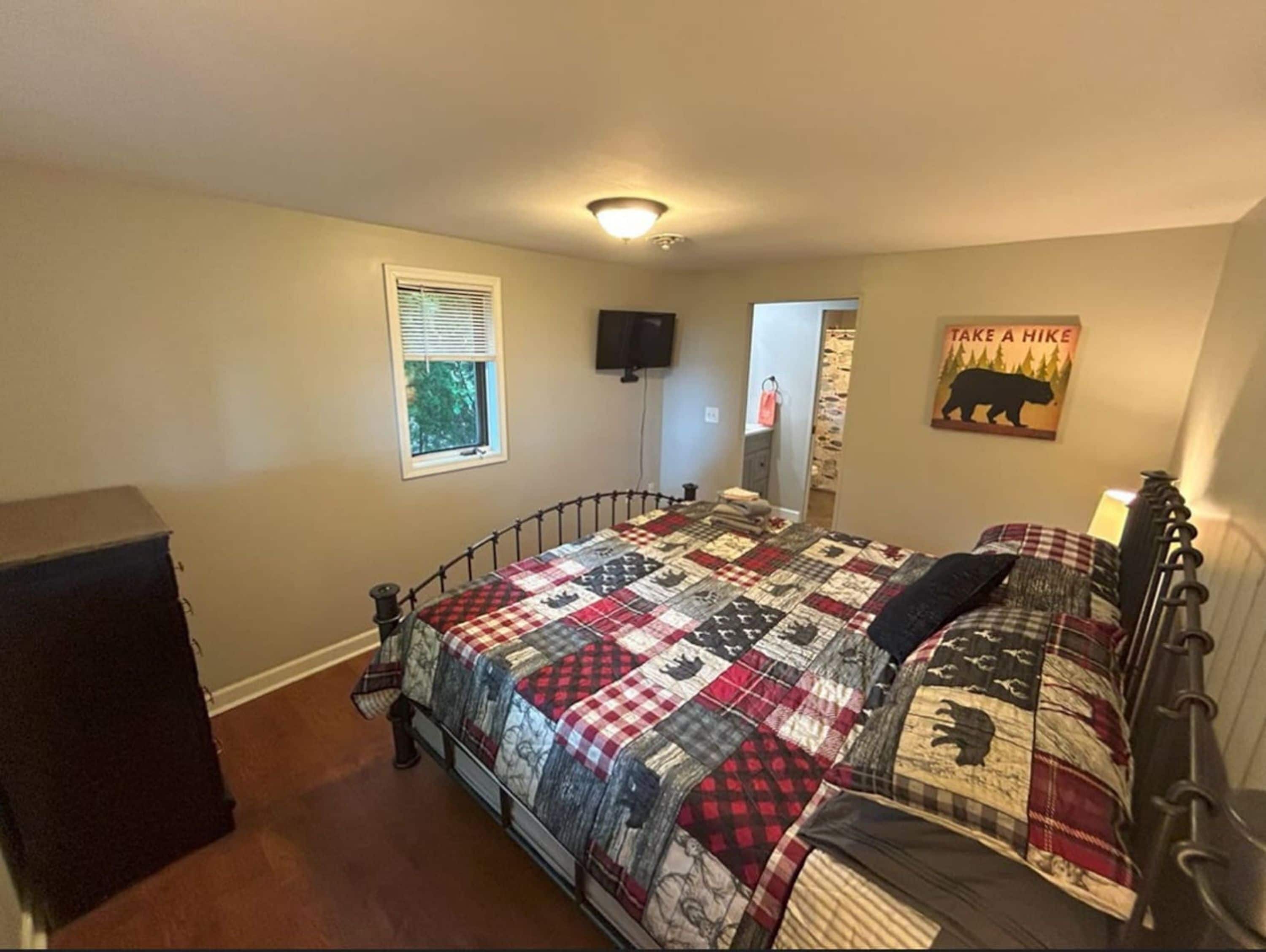 Une chambre à coucher comprenant un lit, une commode, une fenêtre avec des stores, un téléviseur fixé au mur et une affiche.