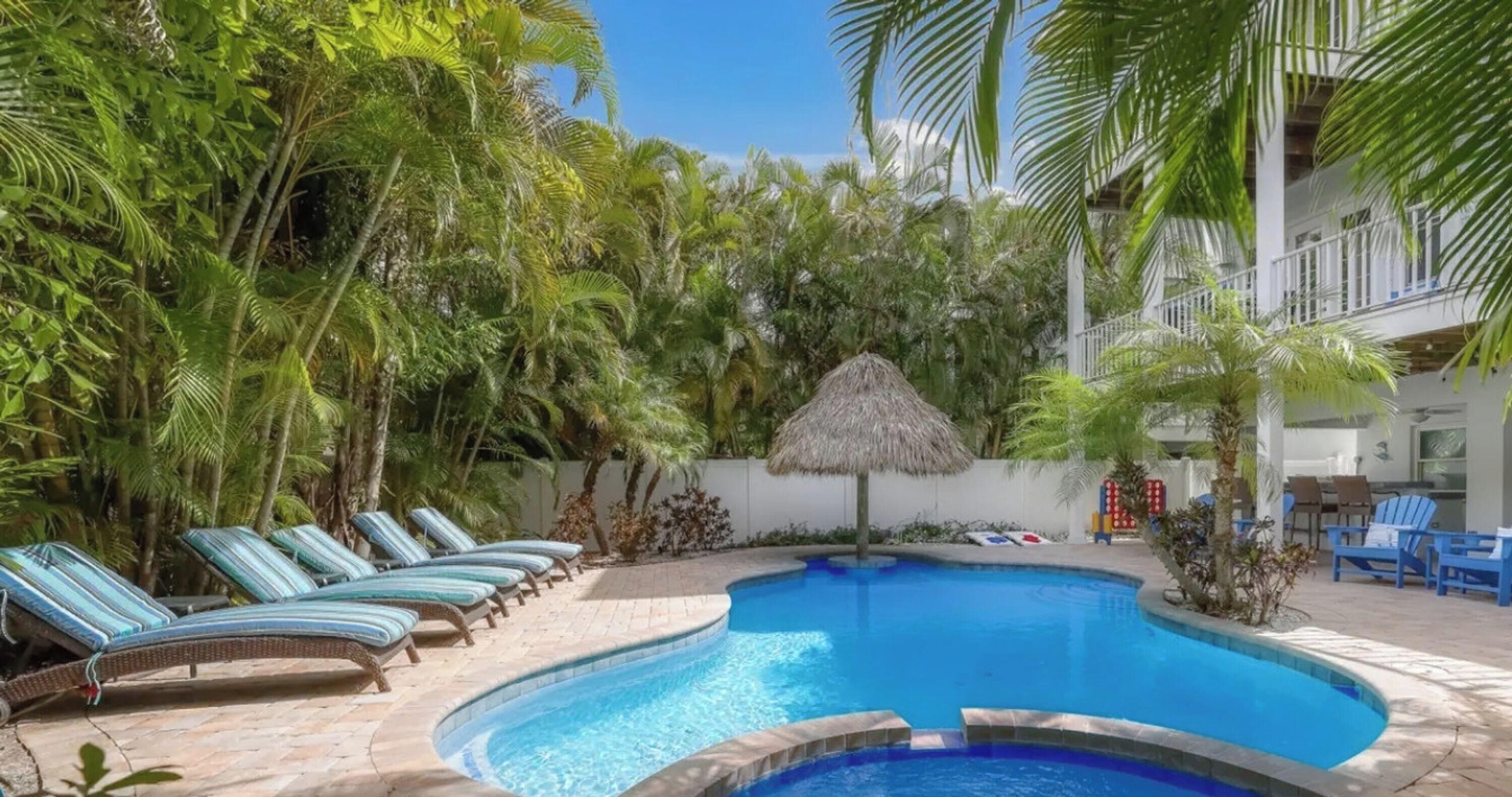 A swimming pool with lounge chairs and a thatched umbrella, surrounded by tropical vegetation.