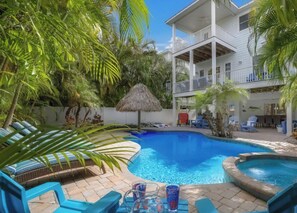 A pool area with lounge chairs, a thatched umbrella, and a building with a balcony. - Fairwinds Tropical Getaway With Pool Hot Tub Game Room Steps to Gulf Beaches (Holmes Beach)