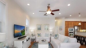 A living room with a ceiling fan, a flat-screen TV, a white sofa, and a kitchen area with wooden cabinets and appliances. - Fairwinds Tropical Getaway With Pool Hot Tub Game Room Steps to Gulf Beaches (Holmes Beach)
