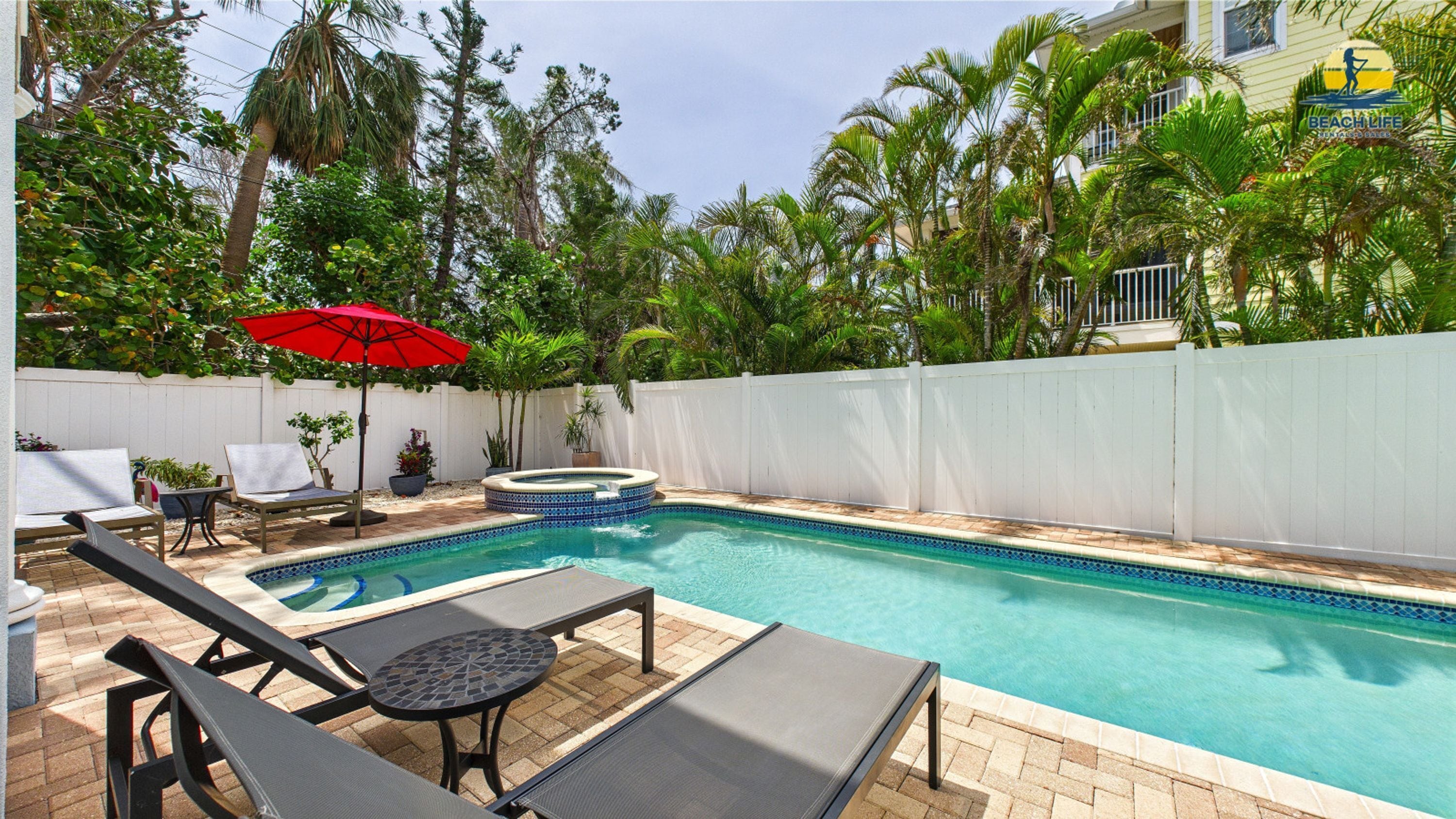 A swimming pool with lounge chairs and a hot tub, surrounded by a white fence and palm trees.