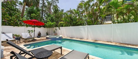 A swimming pool with lounge chairs and a hot tub, surrounded by a white fence and palm trees.