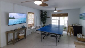 Une pièce équipée d’une table de ping-pong, de ventilateurs de plafond, d’un téléviseur et d’une fenêtre avec des stores.
