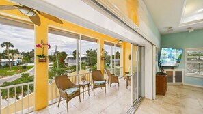 A balcony with wicker chairs, a television, and a view of the neighborhood. - Barefoot Bliss 3-min Walk to Beach Heated Pool Spa Golf Cart Gulf Views (Holmes Beach)