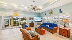 A living room with a blue sofa, wicker furniture, a ceiling fan, and a TV on the wall. - Barefoot Bliss 3-min Walk to Beach Heated Pool Spa Golf Cart Gulf Views (Holmes Beach)