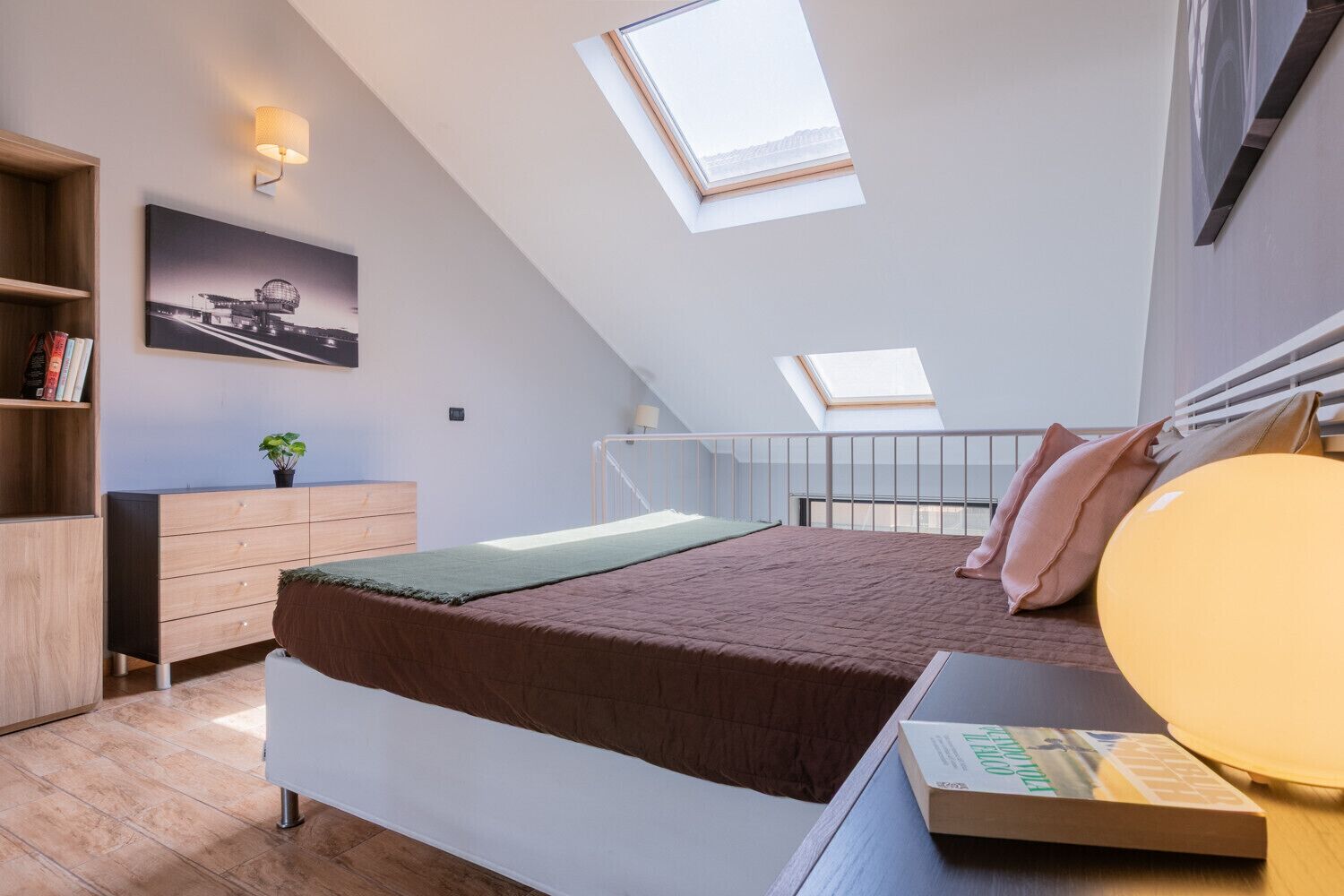 A bedroom with a bed, a bedside table, a lamp, a bookshelf, and a skylight.