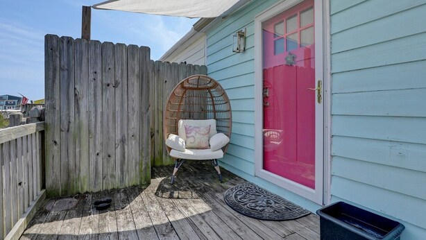 Eine Holzterrasse mit einem Korbstuhl, einer Tür mit einem rosafarbenen Fenster und einem Holzzaun.