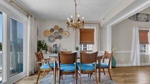 Ein Essbereich mit einer Chandelüster, Holzstühlen mit blauen Kissen, einem gemusterten Teppich und Wanddekoration.