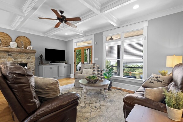 A living room with a stone fireplace, leather furniture, a ceiling fan, and large windows with blinds.