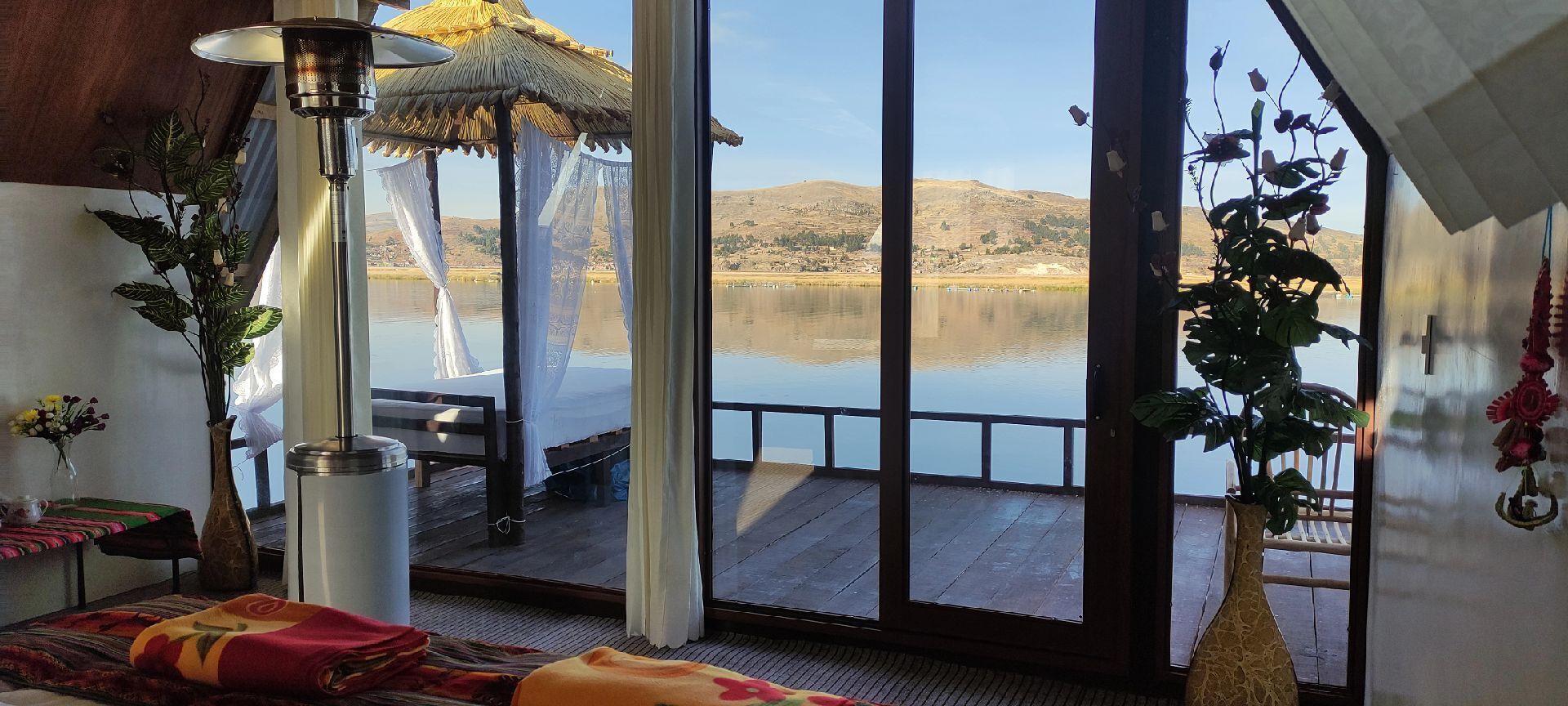 Quarto com uma grande janela com vista para um lago tranquilo, cama com um cobertor colorido, mesa com um vaso de flores e uma planta pendente.
