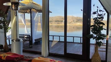 Quarto com uma grande janela com vista para um lago tranquilo, cama com um cobertor colorido, mesa com um vaso de flores e uma planta pendente.