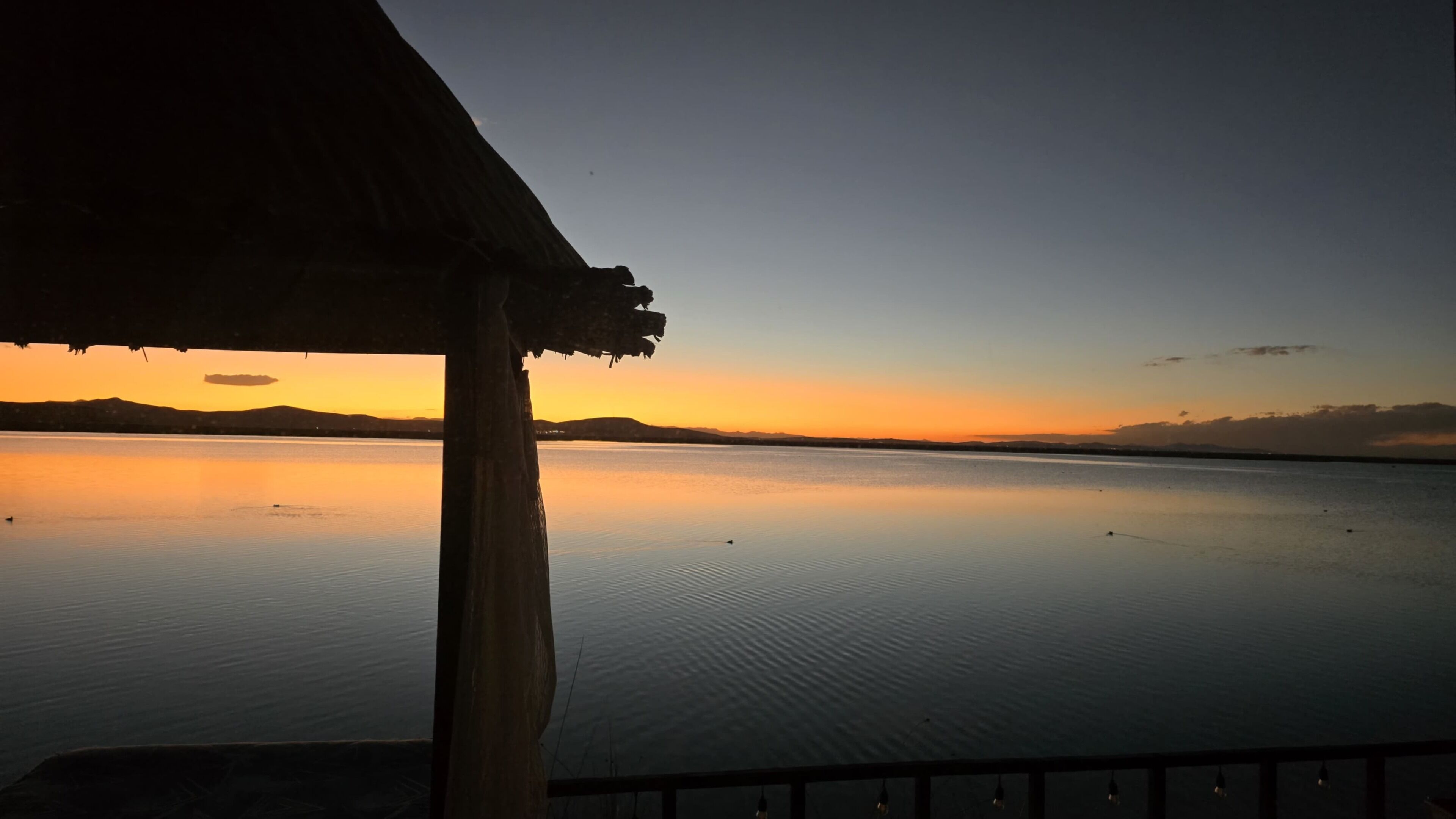 A calm body of water with a sunset in the background, a wooden structure, and a clear sky.