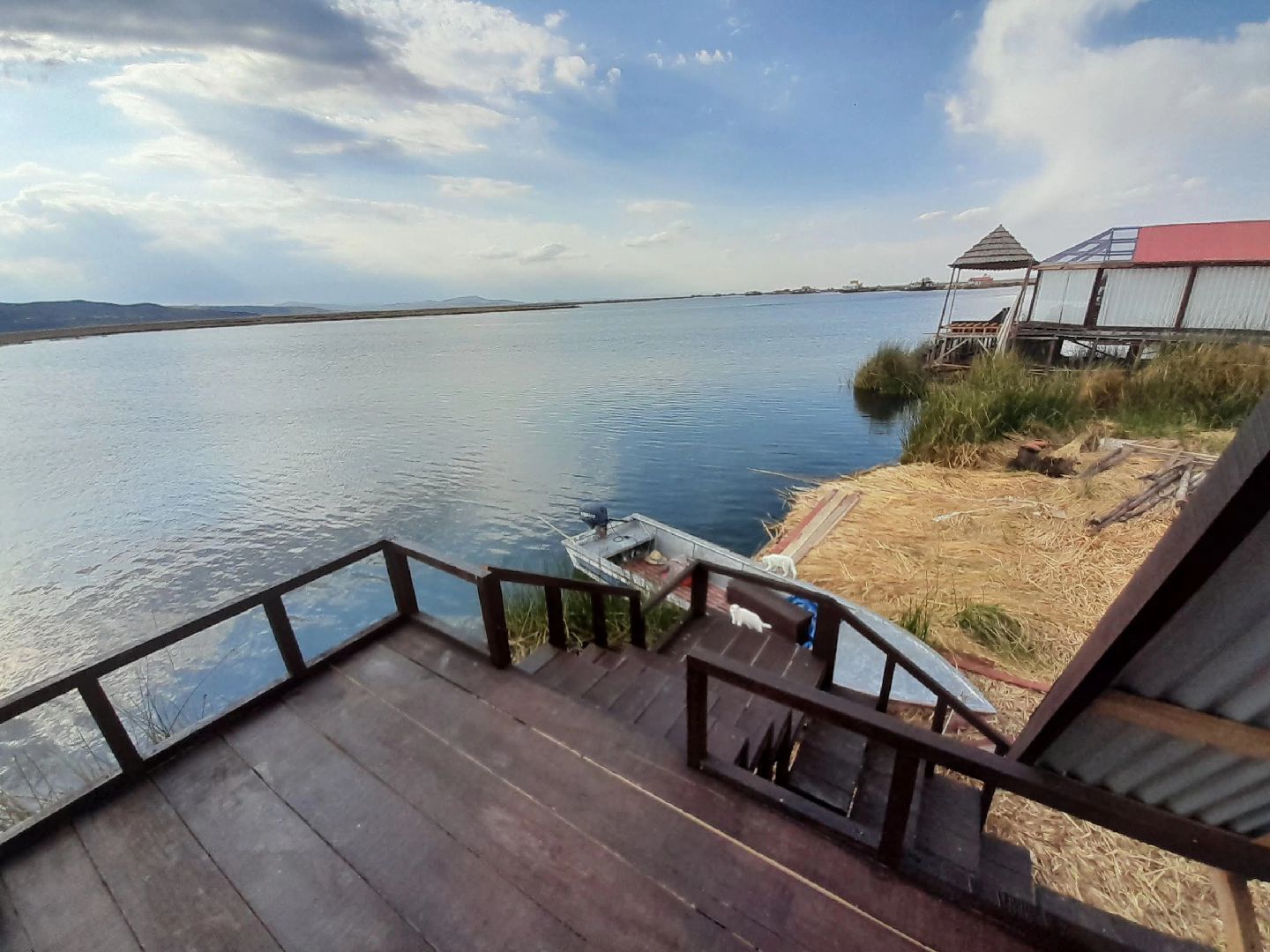 A wooden deck with a railing overlooking a calm body of water, a small dock, and a structure with a red roof.