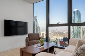 A modern living room with a large TV, comfortable seating, a coffee table with a remote and a plant, and a view of a cityscape through large windows. - Sky-High 1BR, Panoramic City Views with Pool & Gym (Melbourne)