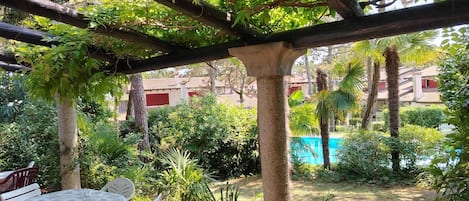 A covered outdoor seating area with a table and chairs, surrounded by lush greenery and a view of a swimming pool.