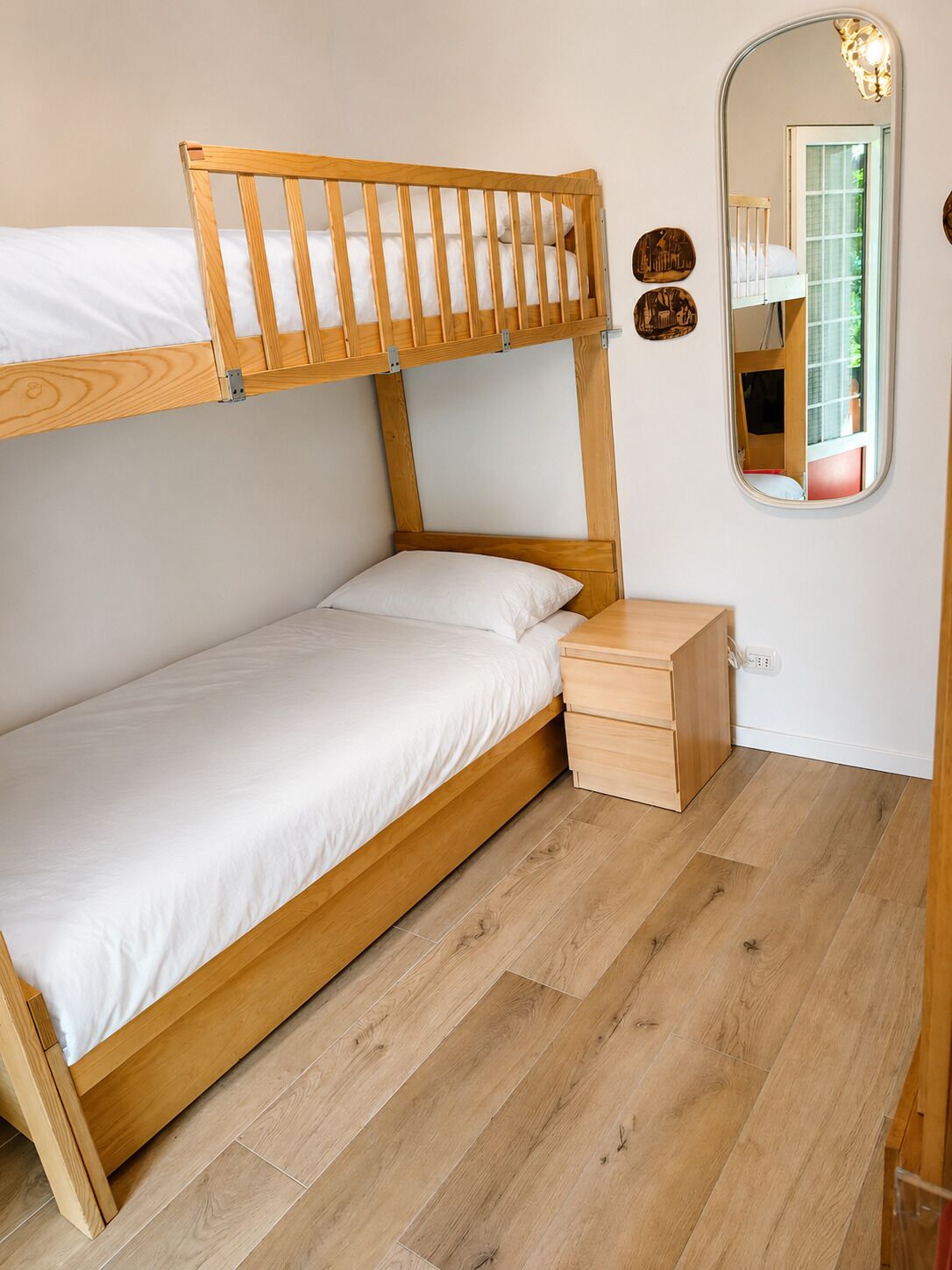A bunk bed with a wooden headboard, a bedside table with drawers, and a mirror on the wall.