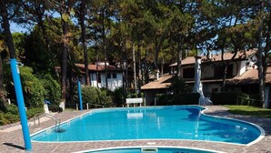 A clear blue swimming pool with a stone border, surrounded by trees and a building with a tiled roof in the background.