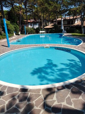 A clear blue swimming pool with a stone border, surrounded by trees and a building with a tiled roof in the background.