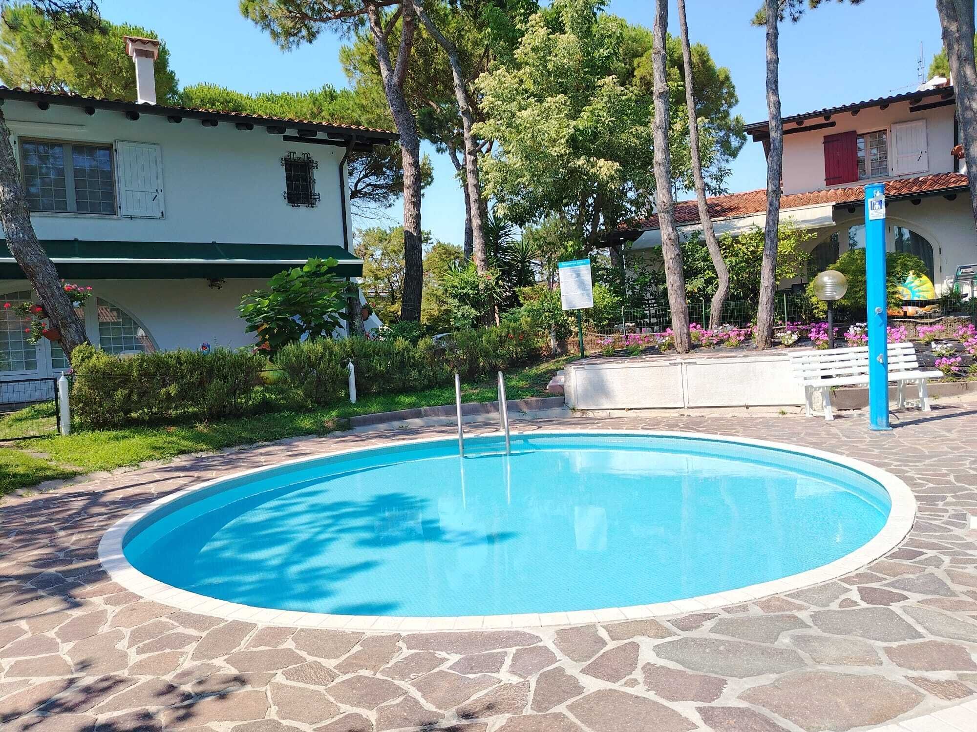A swimming pool with a stone border, surrounded by greenery and a building with a tiled roof.