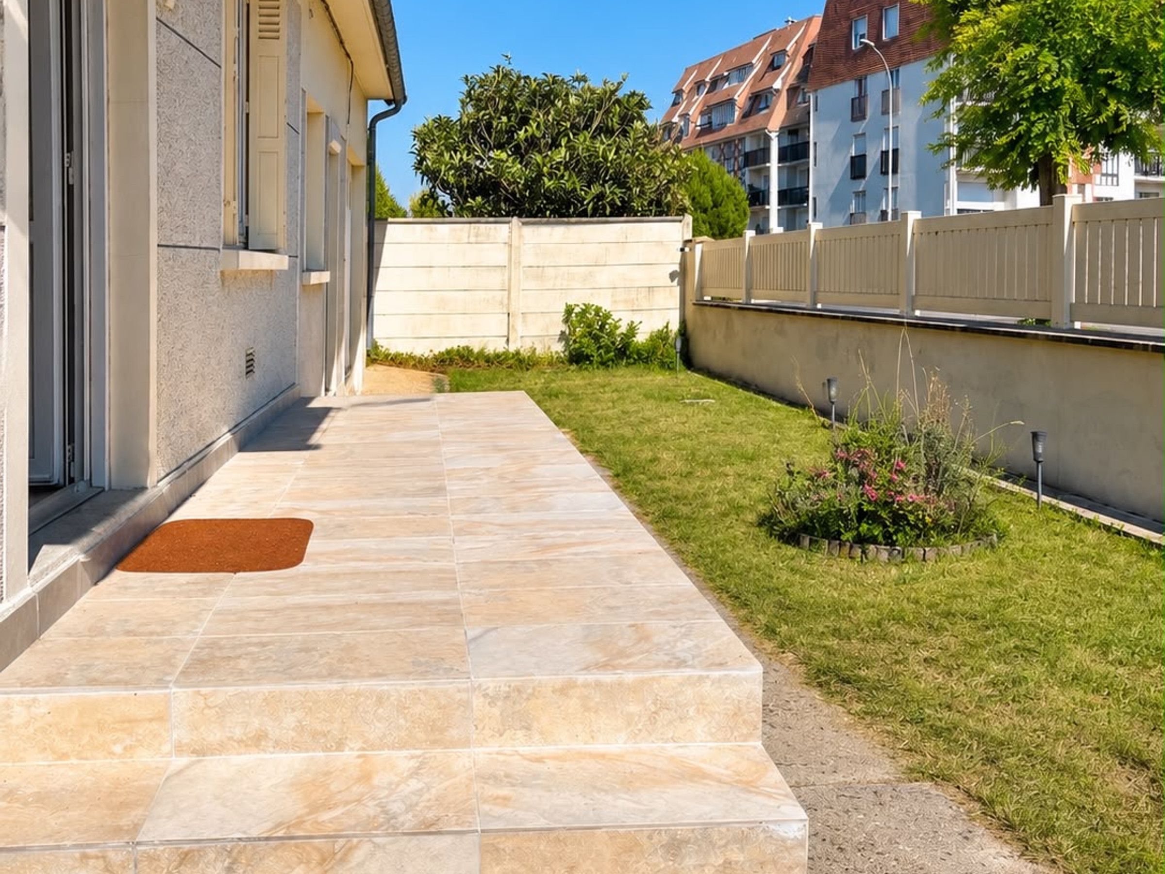 A paved pathway leading to a building entrance, a small grassy area with plants, and a white fence.