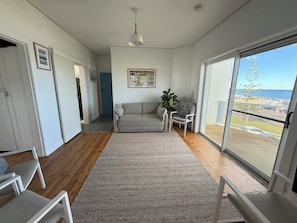 A spacious living area with a grey sofa, a white armchair, a large window with a view of the sea, and a ceiling light. - Alice’s by the Beach, Bar Beach with Beach View, close to town, off street park (Bar Beach)