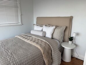 Ein ordentlich bezogenes Bett mit gesteppter Decke, ein Nachttisch mit Lampe und ein Topfpflanze.