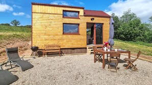 Une cabane en bois avec un toit rouge, deux fenêtres, une porte et une terrasse aménagée avec du mobilier d’extérieur.