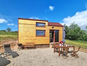 Eine Holzhütte mit rotem Dach, zwei Fenstern, einer Tür und einer Terrasse mit Gartenmöbeln.