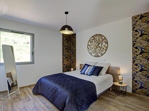 Ein Schlafzimmer mit einem Bett, einem stehenden Spiegel, einer Wanddekoration mit Baummotiv und einem Fenster.