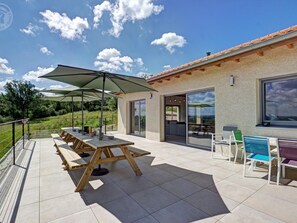 Una terrazza all'aperto con tavoli e sedie in legno, un ombrellone grande e una porta-finestra in vetro che dà su un ambiente interno.